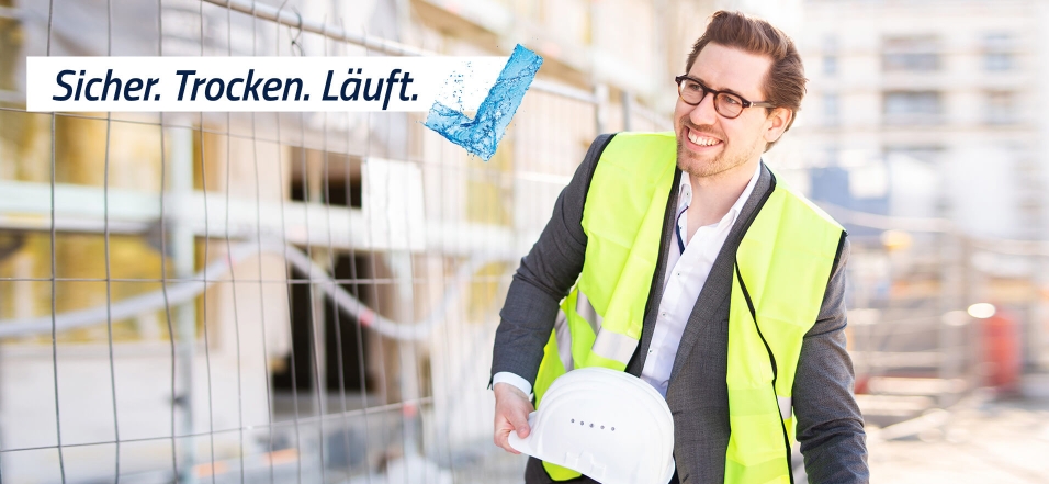 Junger Mann im Anzug mit gelber Warnweste und weißem Sicherheitshelm vor einer Baustelle